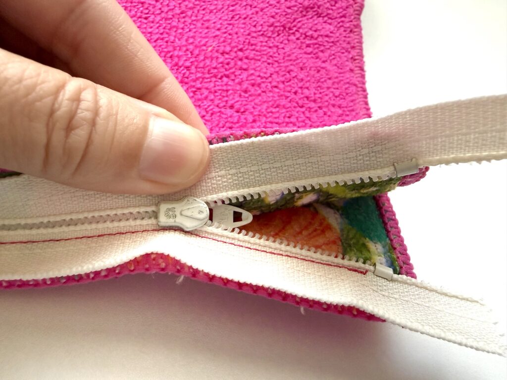 A hand holding open a white zipper on a pink fabric pouch, revealing a multicolored fabric lining inside.