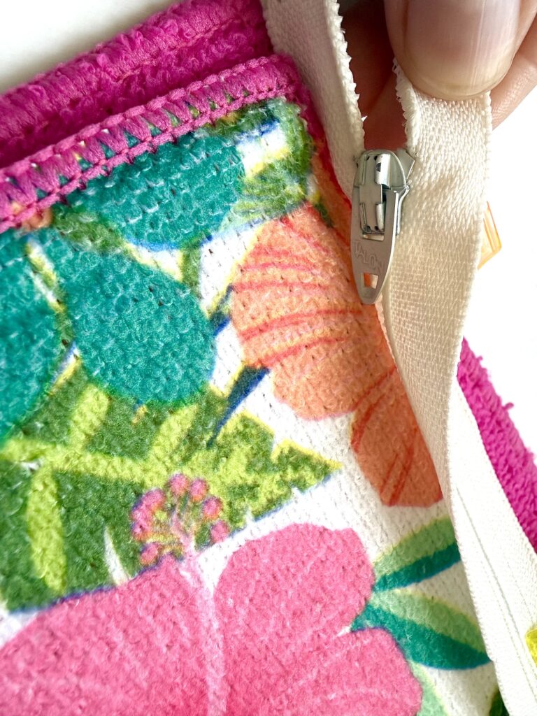 A hand holds a white zipper on a colorful floral fabric with pink, orange, and green patterns and pink stitched edging.