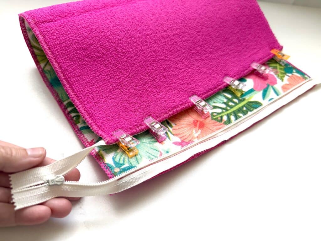 A hand holds a white zipper next to a pink fabric pouch with floral lining, secured with clips along the edge.
