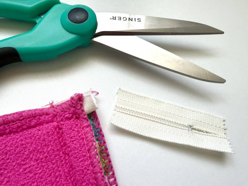 A pair of teal scissors, a pink fabric piece with stitching, and a short white zipper segment are arranged on a white surface.