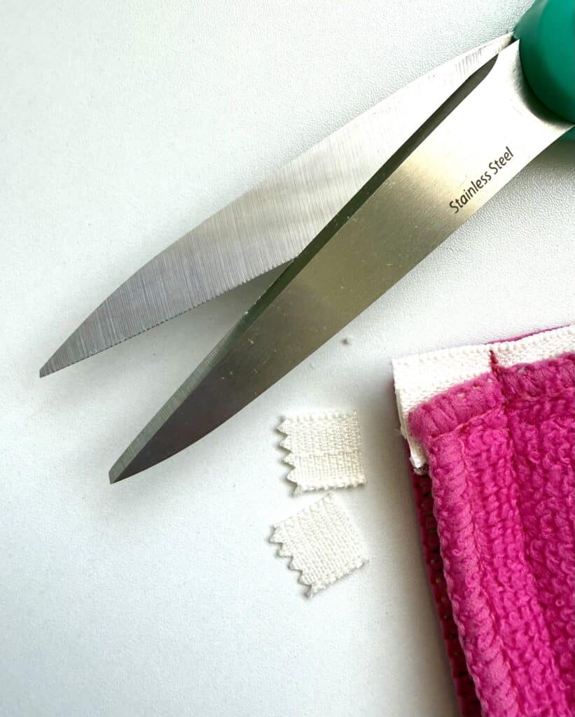 Large stainless steel scissors cutting a textured white fabric, with small pieces on a white surface next to a pink cloth.