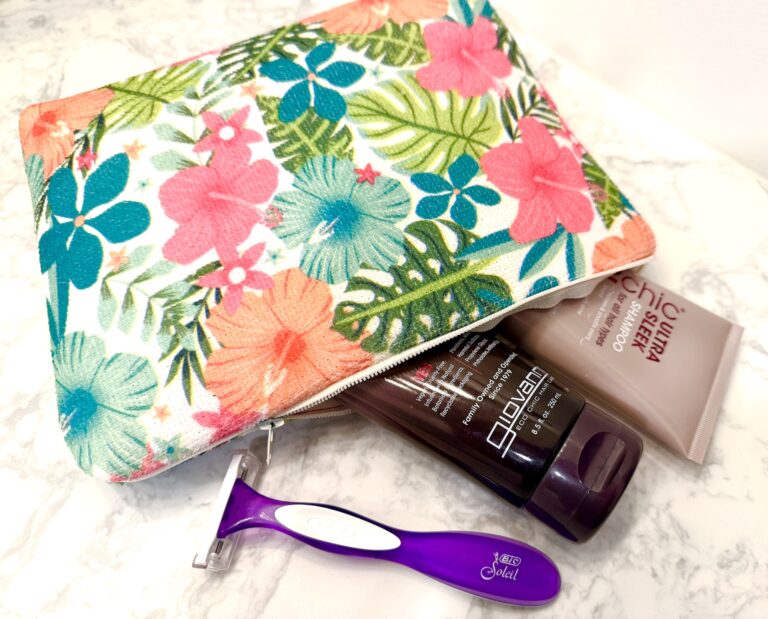 A floral cosmetic bag with a razor, a brown bottle of body wash, and a tube of body lotion on a marble surface.