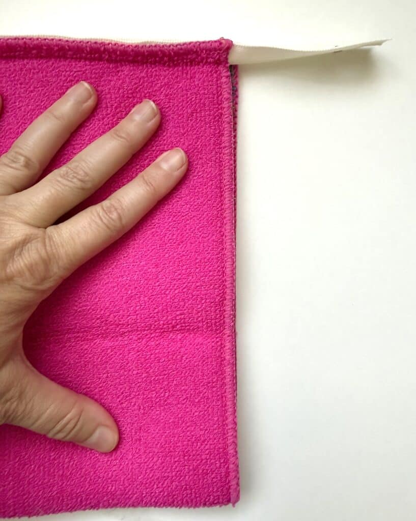 A left hand rests on a folded bright pink cloth with a white tag sticking out on the top right, all on a white surface.