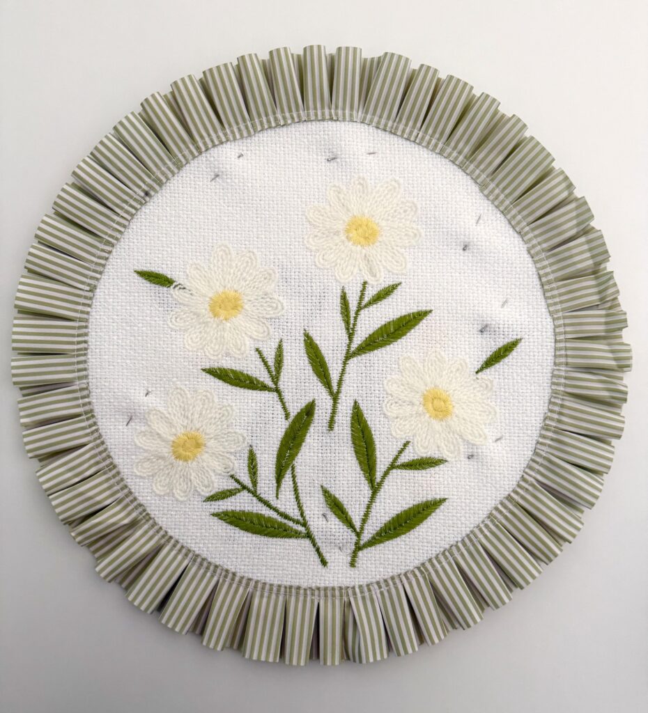 two circular placemats with embroidered daisies and green and white striped pleated edge pinned together and displayed on a white background
