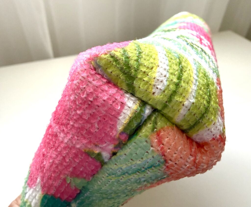 A close-up of a colorful, textured object with pink, green, orange, and white patterns, possibly a cushion or fabric item, shown against a light background.