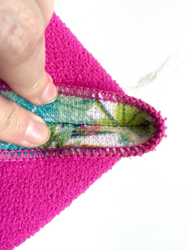 A hand pulls back the edge of a bright pink fabric, revealing a colorful, patterned inner lining against a white background.