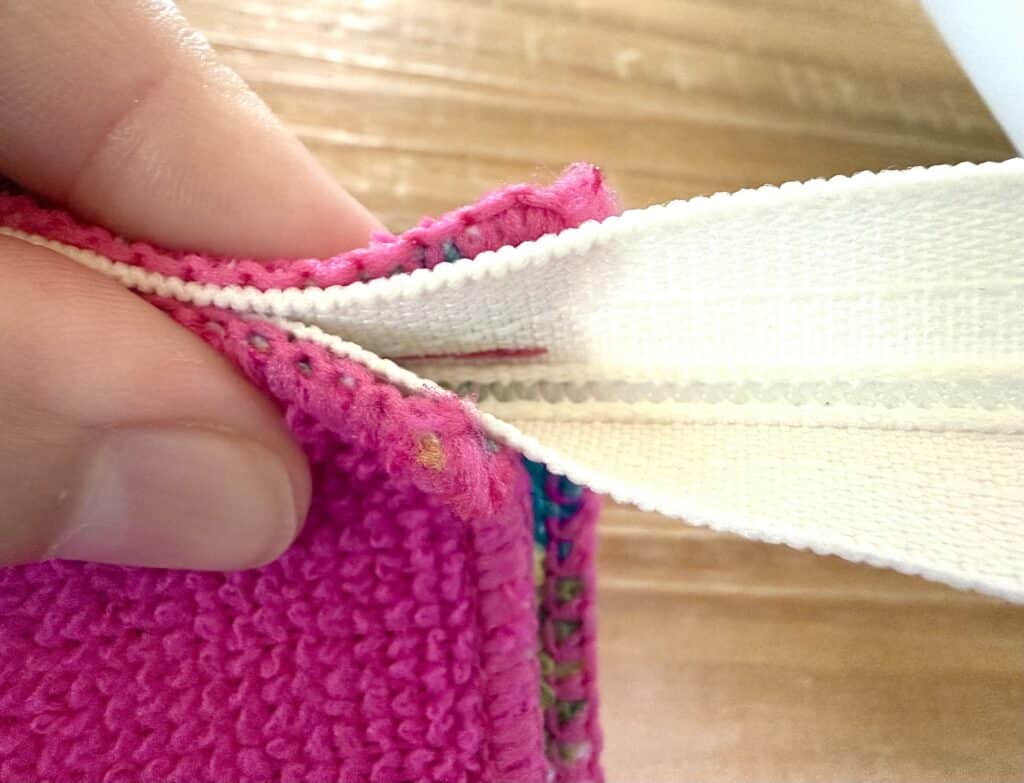 Close-up of a hand holding a pink fabric with a white zipper partially unzipped, showing the stitching details and fabric texture.