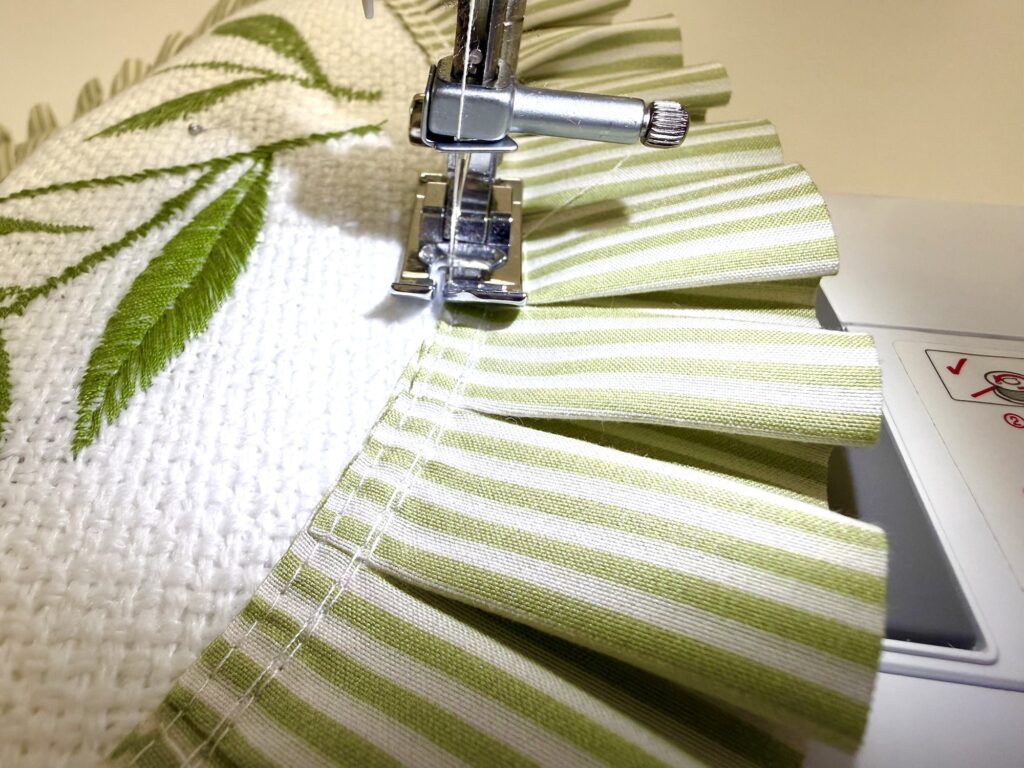 sewing machine needle inserted into the white textured fabric of a placemat pillow with green and white striped ruffles on the other side of the needle