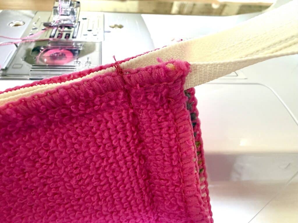 Close-up of a sewing machine stitching a white strap to a bright pink textured fabric, showing the needle and presser foot in action.