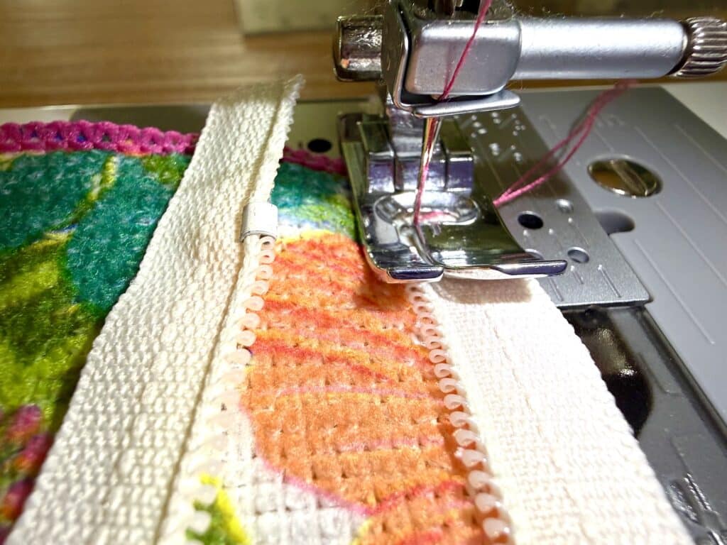 Close-up of a sewing machine needle stitching colorful fabric with a zipper and white trim under the presser foot.