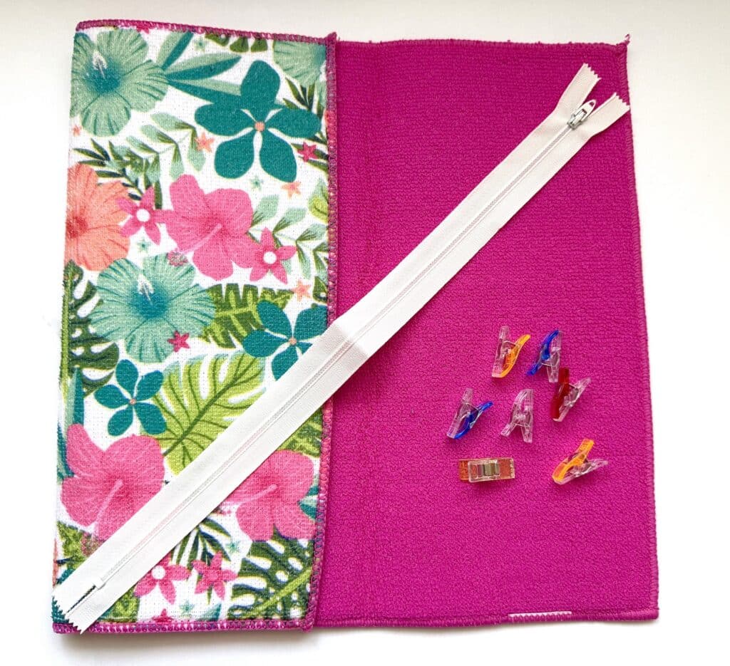 Two pieces of fabric, a white zipper, and six colorful sewing clips are arranged on a white background.