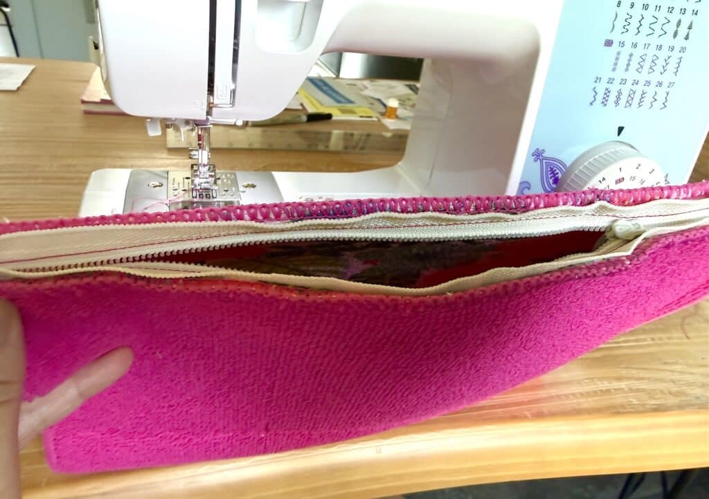 A hand holds open a bright pink zippered pouch in front of a sewing machine on a wooden table.