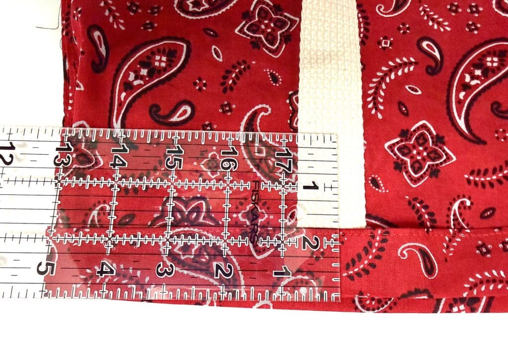 Clear ruler placed on red bandana fabric with white and black paisley patterns, measuring the material’s width and showing inches and centimeters on the grid.