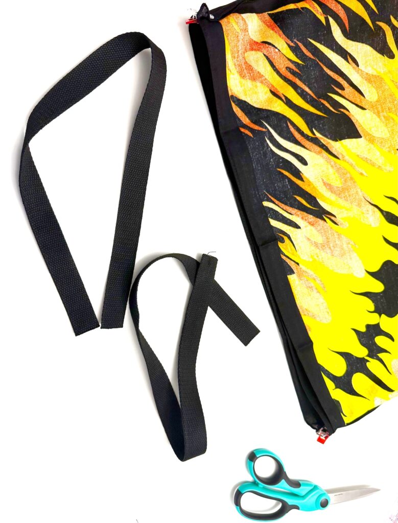 A cut black strap, a pair of teal scissors, and a vibrant bandana bag with a yellow and orange flame pattern are displayed on a white surface.