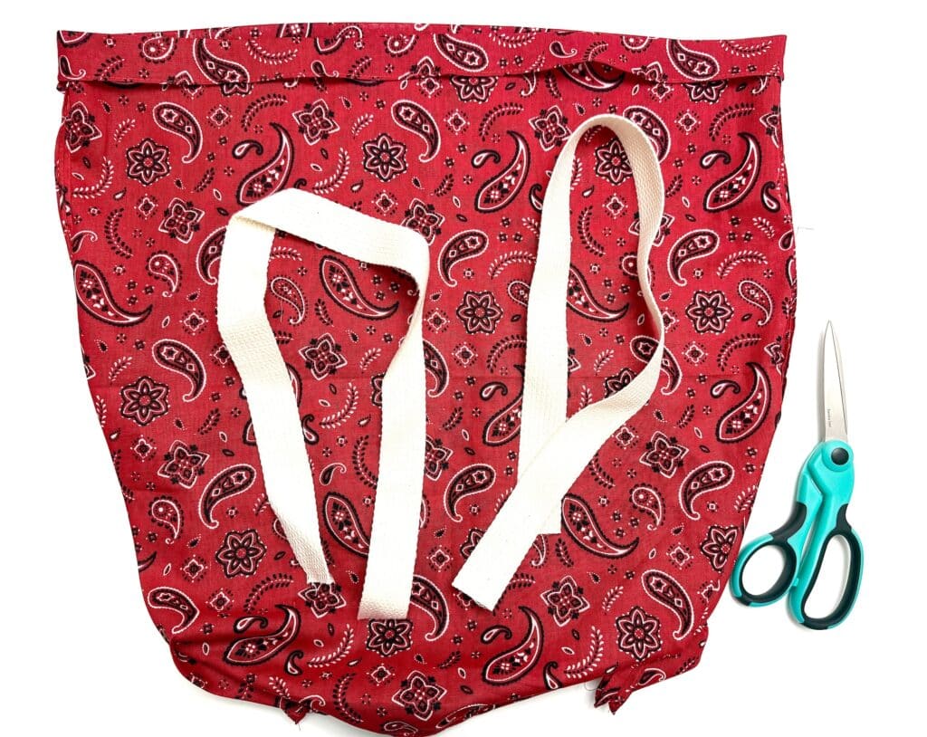 A red bandana-patterned fabric bag with white straps is next to a pair of teal-handled scissors on a white background.