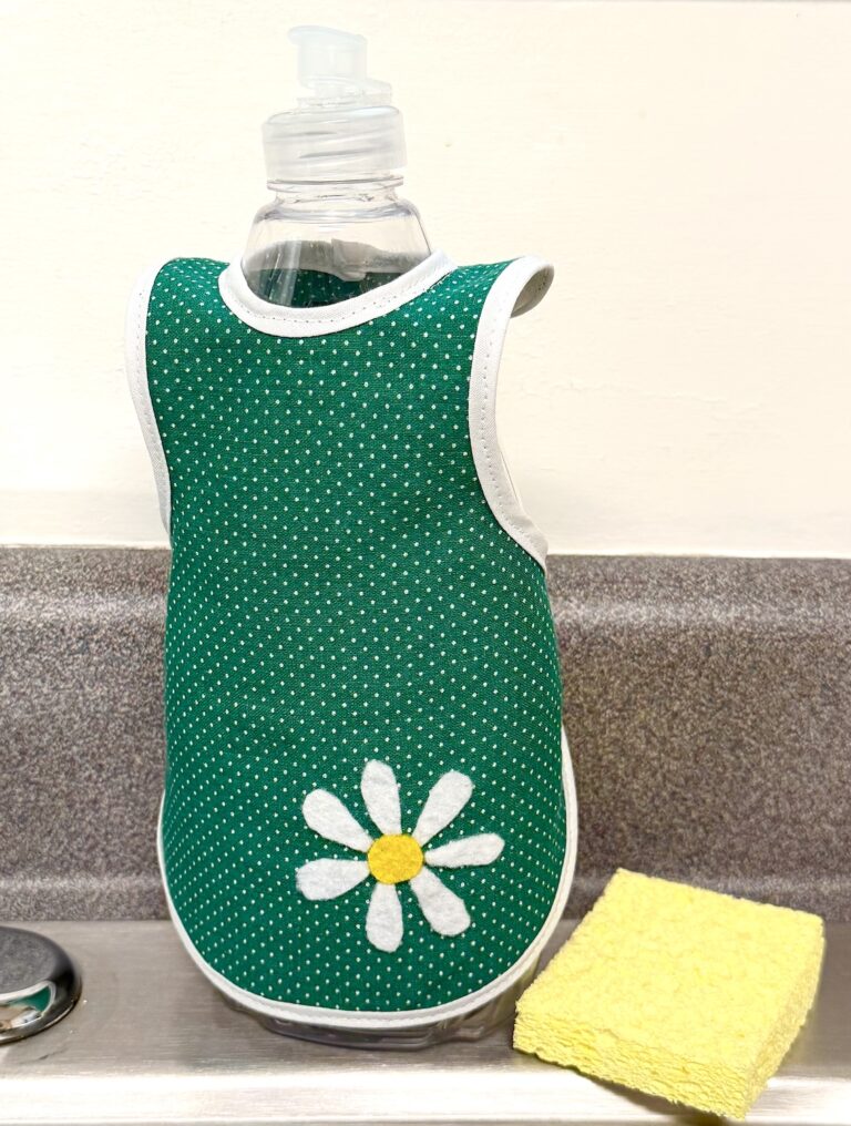 green fabric apron with a daisy appliqué covering a dish soap bottle at the kitchen sink with a yellow sponge nearby