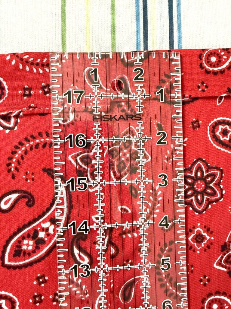 A clear ruler placed vertically on a red paisley fabric and white cloth with green and blue stripes, showing measurements from 13 to 18 inches.