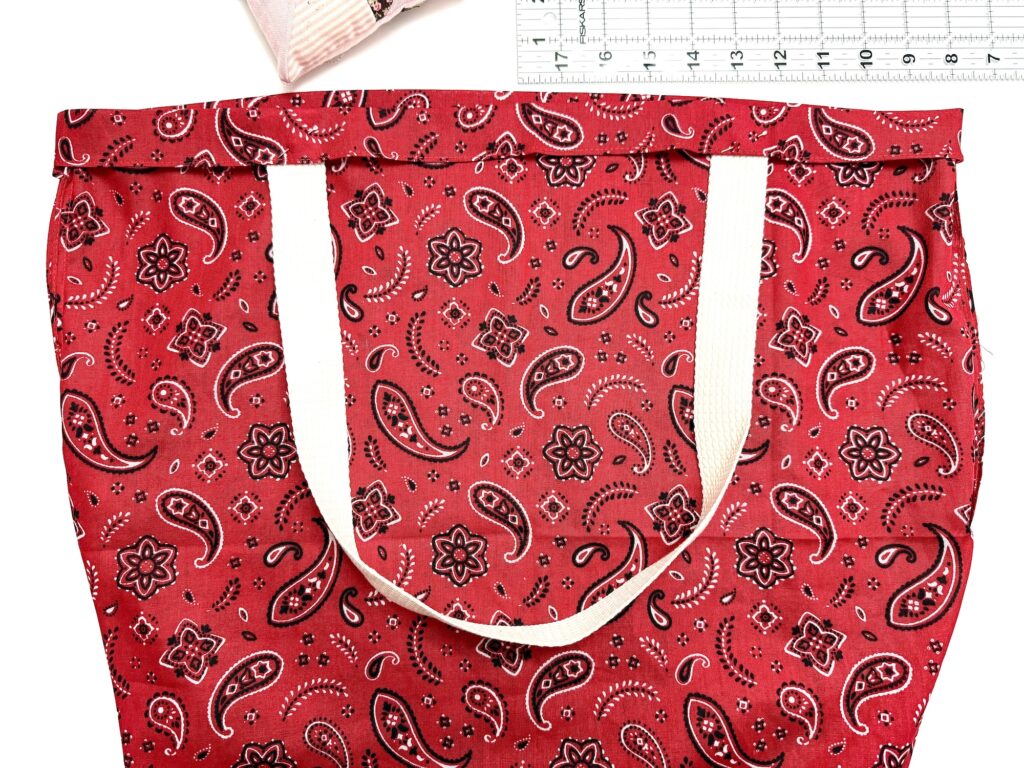 Red tote bag with a white strap and paisley pattern, lying flat next to a ruler and a small striped pouch in the corner.