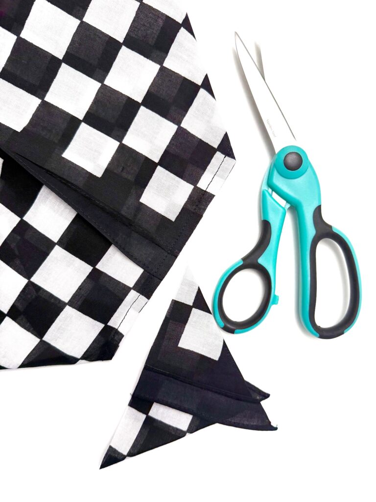A pair of blue scissors and black-and-white checkered fabric, perfect for crafting a stylish bandana bag, with two triangular pieces cut out are shown on a white background.
