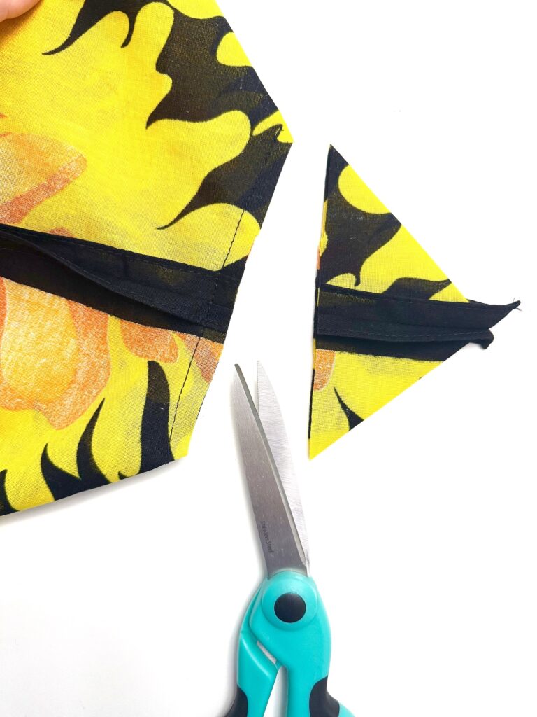 A pair of teal-handled scissors cutting a triangular piece from yellow and black flame-patterned fabric—perfect for crafting a unique bandana bag—on a white background.