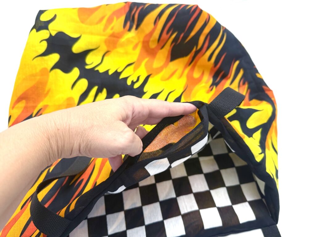 A hand opens a bandana bag with a yellow and orange flame pattern outside and black-and-white checkerboard lining inside.