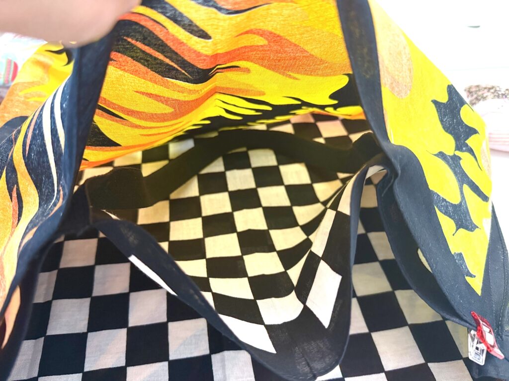 A hand holds open a colorful bandana bag with a yellow and orange pattern and a black-and-white checkered lining, above a matching checkered surface.