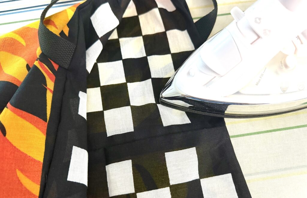 A steam iron rests on a black and white checkered cloth with a flame pattern, both laid on an ironing board, beside a stylish bandana bag.