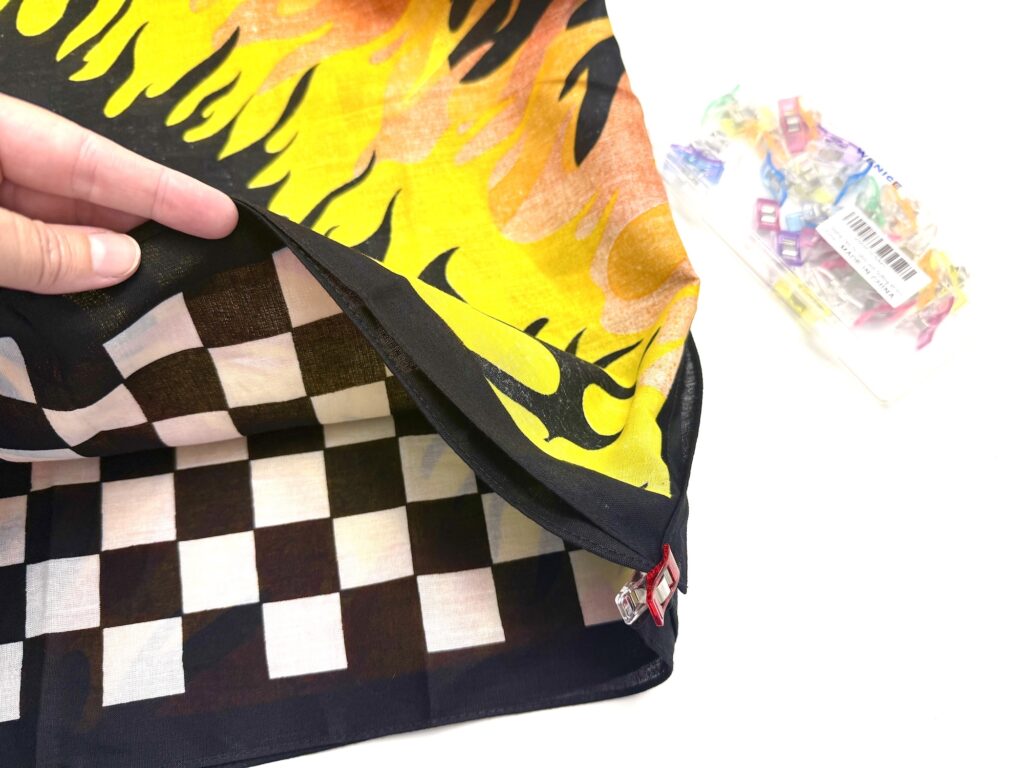 A hand holds layered fabric with a flame and checkerboard pattern, secured by sewing clips—perfect for crafting your own unique bandana bag. Colorful clips are scattered on the side.