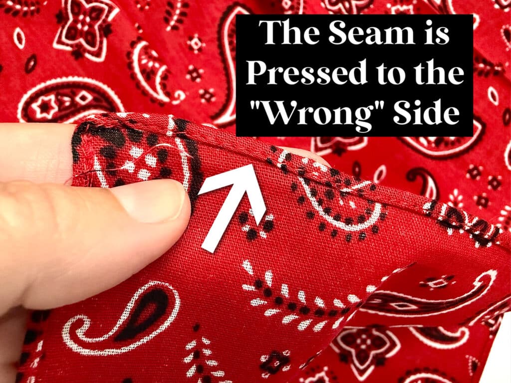 A hand holds red patterned fabric with a seam pressed to the back side. An arrow points to the seam, and text reads: “The Seam is Pressed to the ‘Wrong’ Side.”.