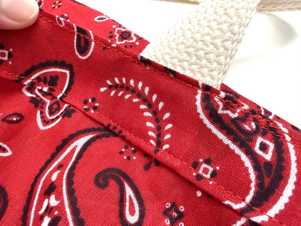 Close-up of a red bandana-patterned fabric tote bag with a cream-colored woven handle and visible stitching details.