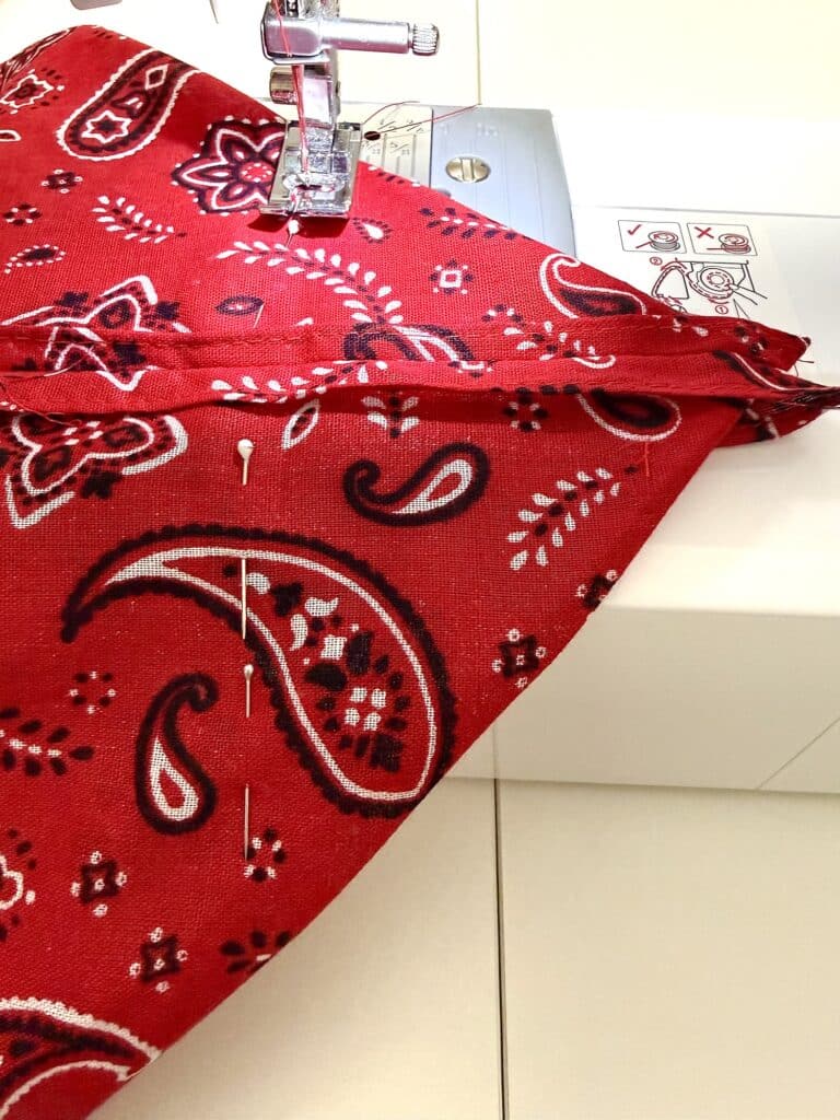 Red bandana fabric with pins is being sewn on a white sewing machine, showing the presser foot, needle, and part of the machine’s control panel.