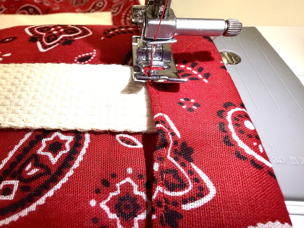 Close-up of a sewing machine stitching red paisley fabric with a cream textured strap underneath the presser foot.