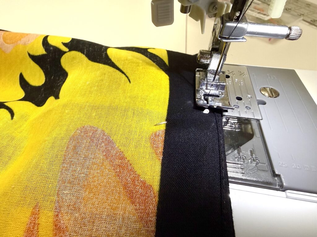 A sewing machine stitching a black seam on a piece of yellow and orange printed fabric, with a pin holding the fabric in place—perfect for crafting a unique bandana bag.