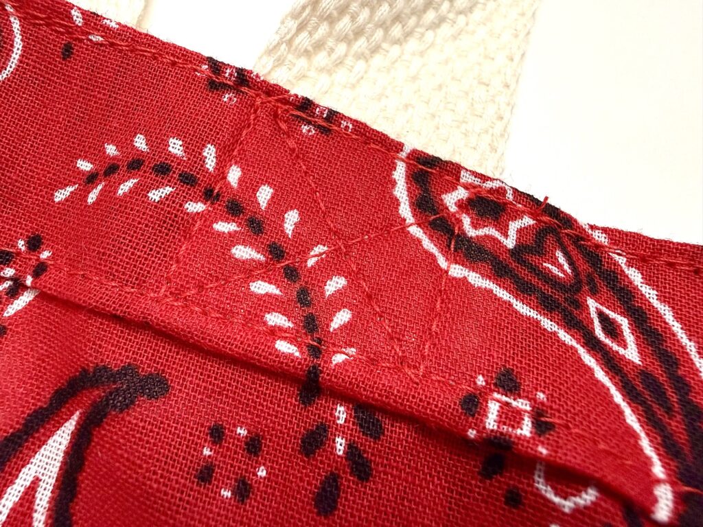 Close-up of a red fabric with a bandana pattern, white and black designs, and visible stitching securing a cream-colored textured strap.
