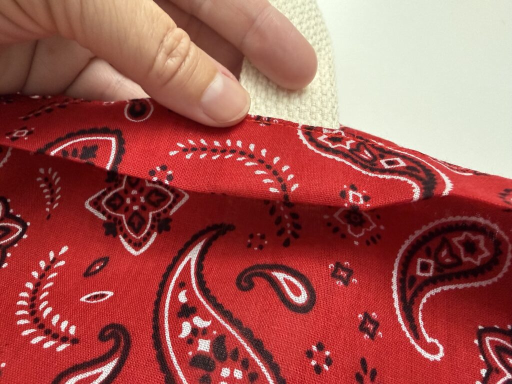 A hand holds up a piece of red fabric with a paisley pattern, revealing a layer of beige textured fabric underneath.