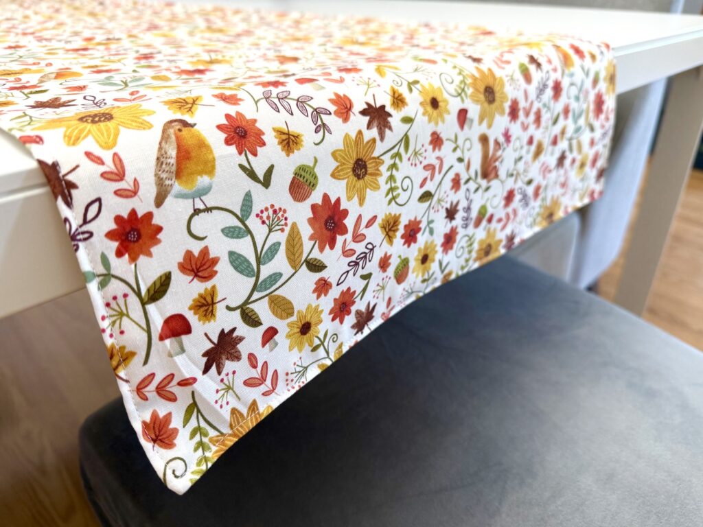 A table runner patterned with autumn leaves, flowers, acorns, and a small bird, draped over a white table.