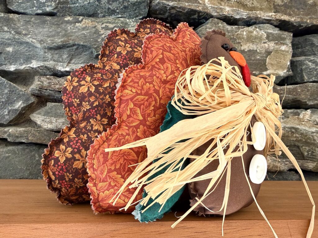 Fabric turkey decoration with patterned leaf-print tail feathers, a raffia bow around its neck, and white button accents on a stone background.