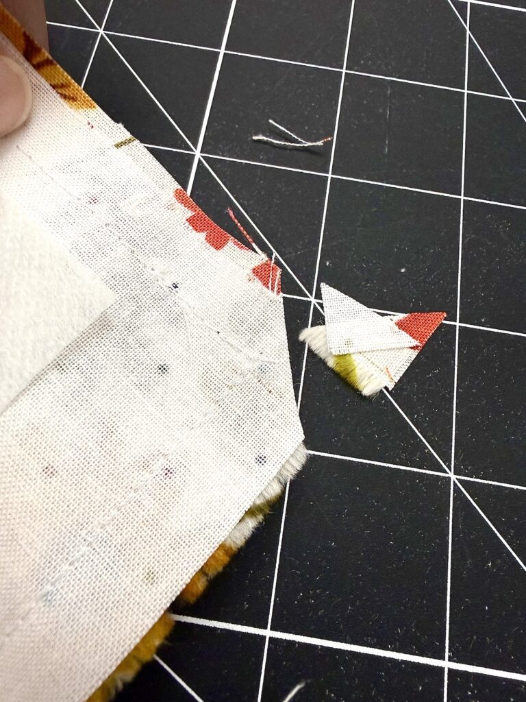 A corner of fabric with a sewn seam resting on a cutting mat, with a small triangular fabric scrap trimmed off and sitting nearby.