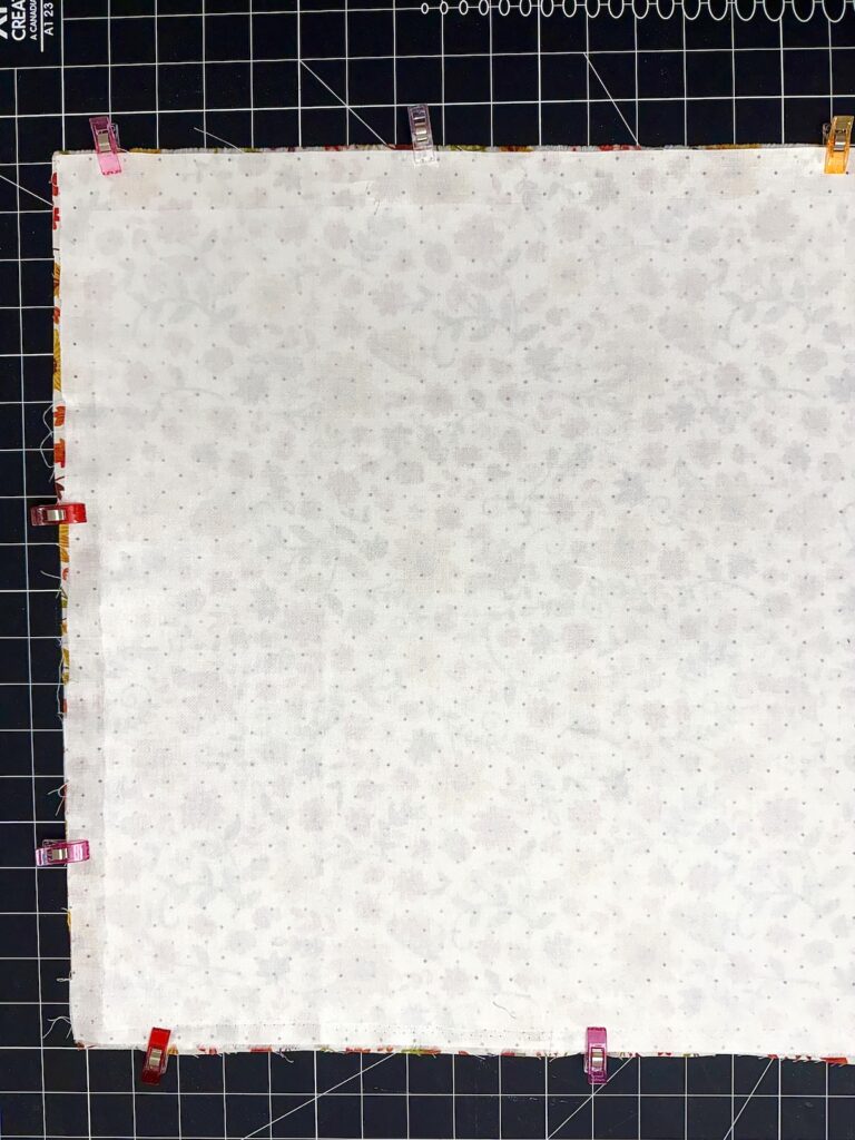 A square piece of light-colored floral fabric secured with sewing clips to a black cutting mat.