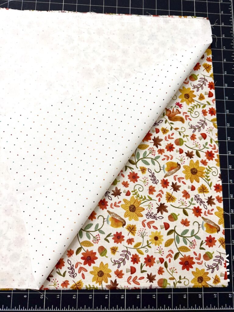 Two layers of fabric on a cutting mat: one white with multicolored dots, and one with a floral and woodland animal print.