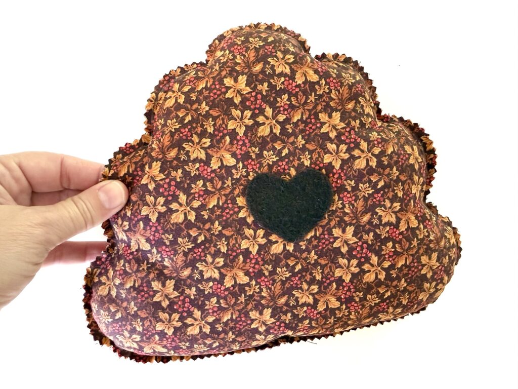 A hand holds a cloud-shaped fabric pillow patterned with autumn leaves and berries, featuring a small black felt heart in the center.