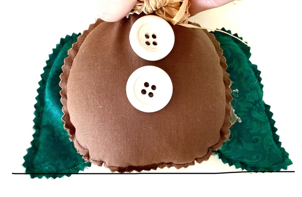 A brown, circular fabric ornament with two white buttons on the front and green fabric wings on the sides.