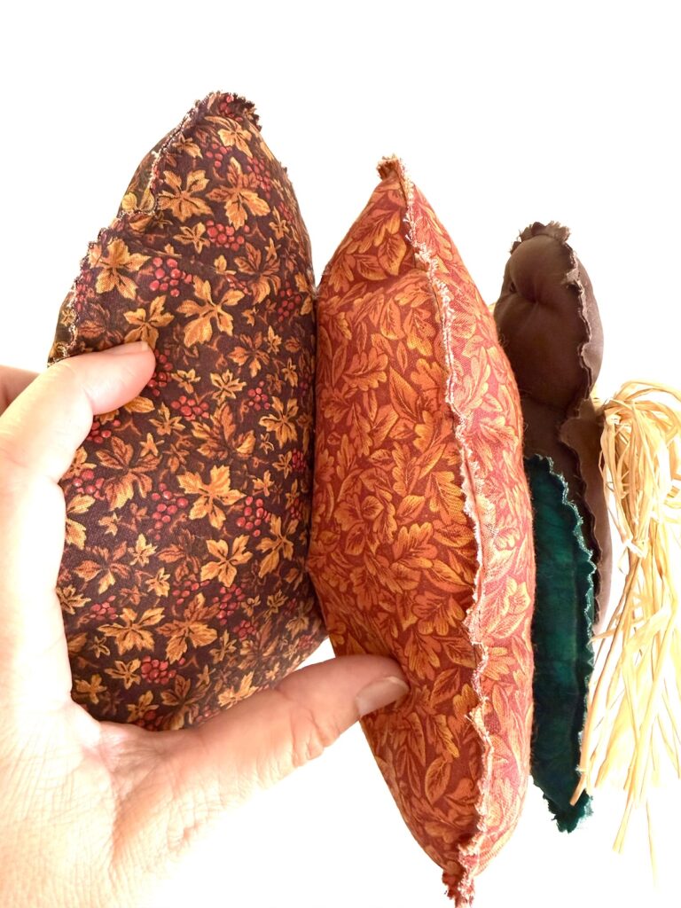 A hand holds three fabric plush shapes: two with orange and brown autumn leaf patterns and one dark brown shape with raffia trim.