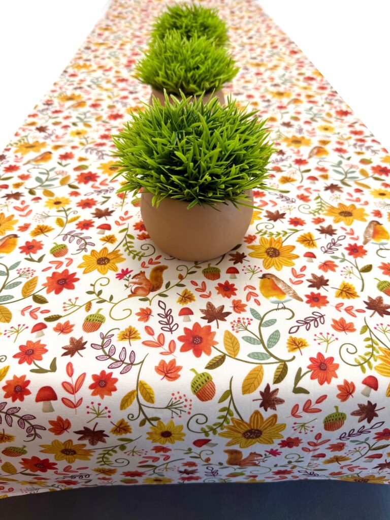 Three small artificial grass plants in pots centered on a table runner patterned with colorful autumnal flowers, squirrels, and birds.
