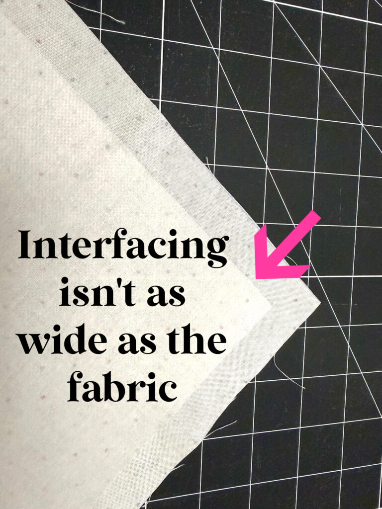 A piece of fabric with a smaller piece of interfacing on top, with a pink arrow pointing to the edge and the text: “Interfacing isn’t as wide as the fabric.”