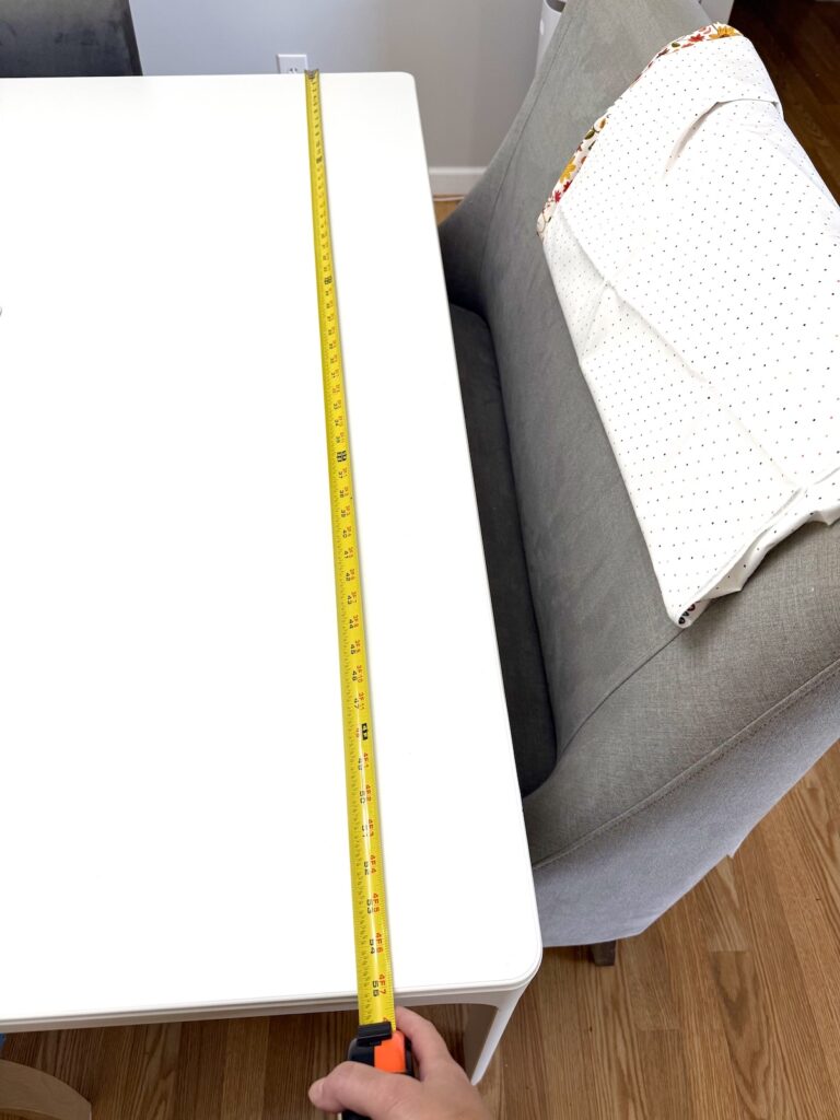 A hand uses a yellow measuring tape to measure the length of a white rectangular table next to a gray upholstered bench.