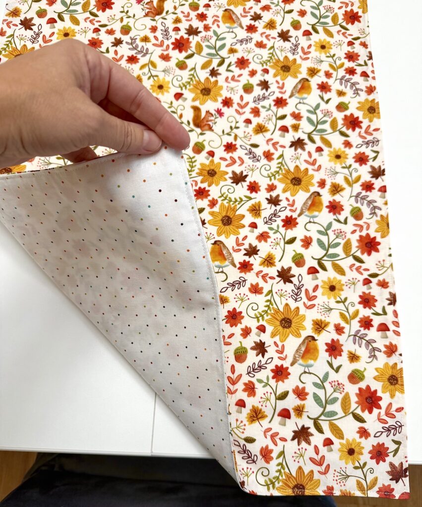 A hand lifts the corner of a double-sided table runner, revealing a white side with multicolored dots and an autumn-themed side with flowers, leaves, birds, and acorns.