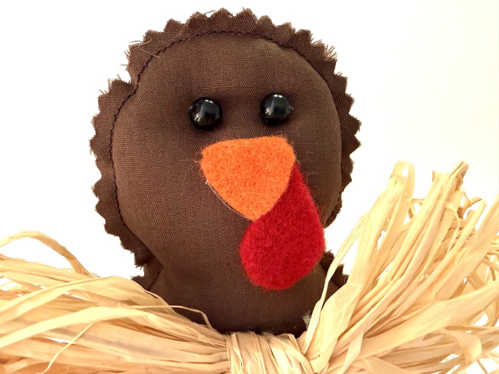 A brown fabric turkey ornament with black bead eyes, an orange beak, and a red wattle, nestled in dried raffia.