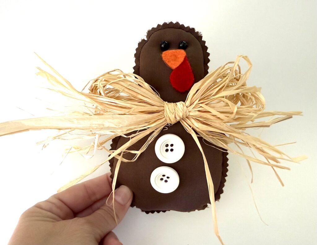 A brown felt turkey ornament with a raffia bow and two white button embellishments, held by a hand.