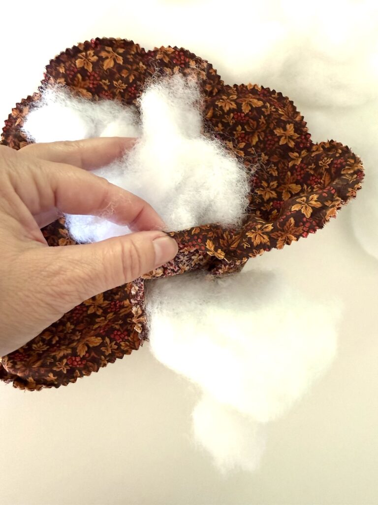 A hand stuffs fluffy white polyester fiberfill into a brown fabric casing decorated with a pattern of autumn leaves and berries.